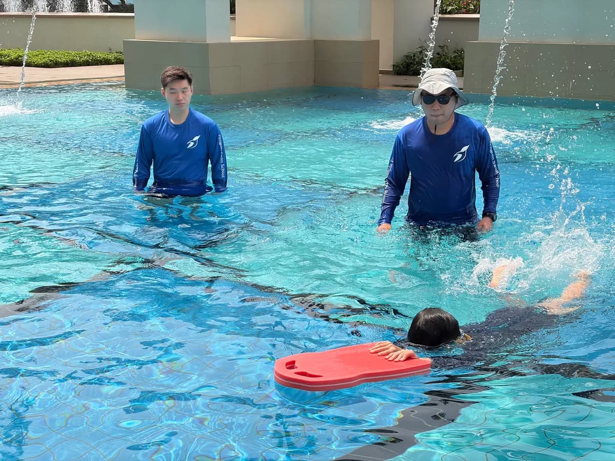 Coach Rayner wearing a hat and sunglasses mentoring a new coach during a Penguin Swim School lesson as a learner swims nearby